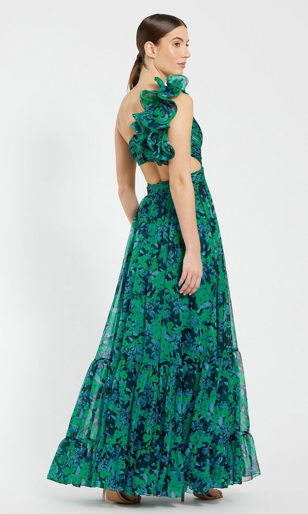 A woman stands sideways wearing the Mac Duggal 68650 One-Shoulder Print Formal Dress, a sleeveless, floor-length green and blue floral gown with ruffled shoulder detail.